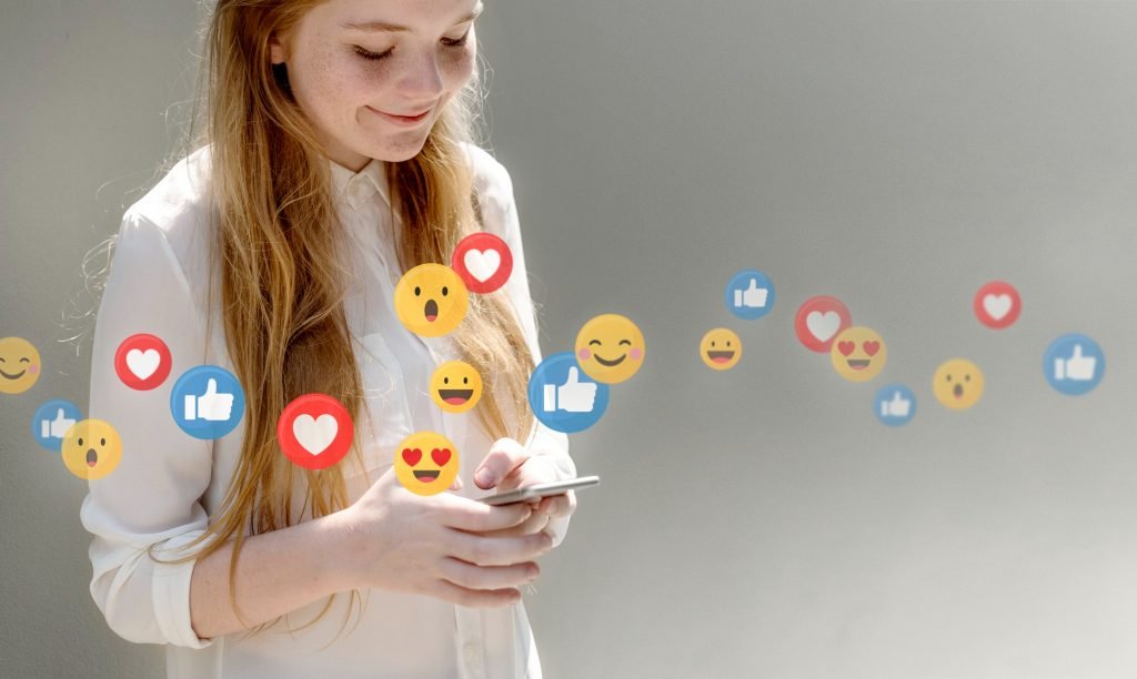 Mujer joven sonriendo y viendo dando like su celular, y se ven iconos de like y emoticones saliendo de la pantalla.